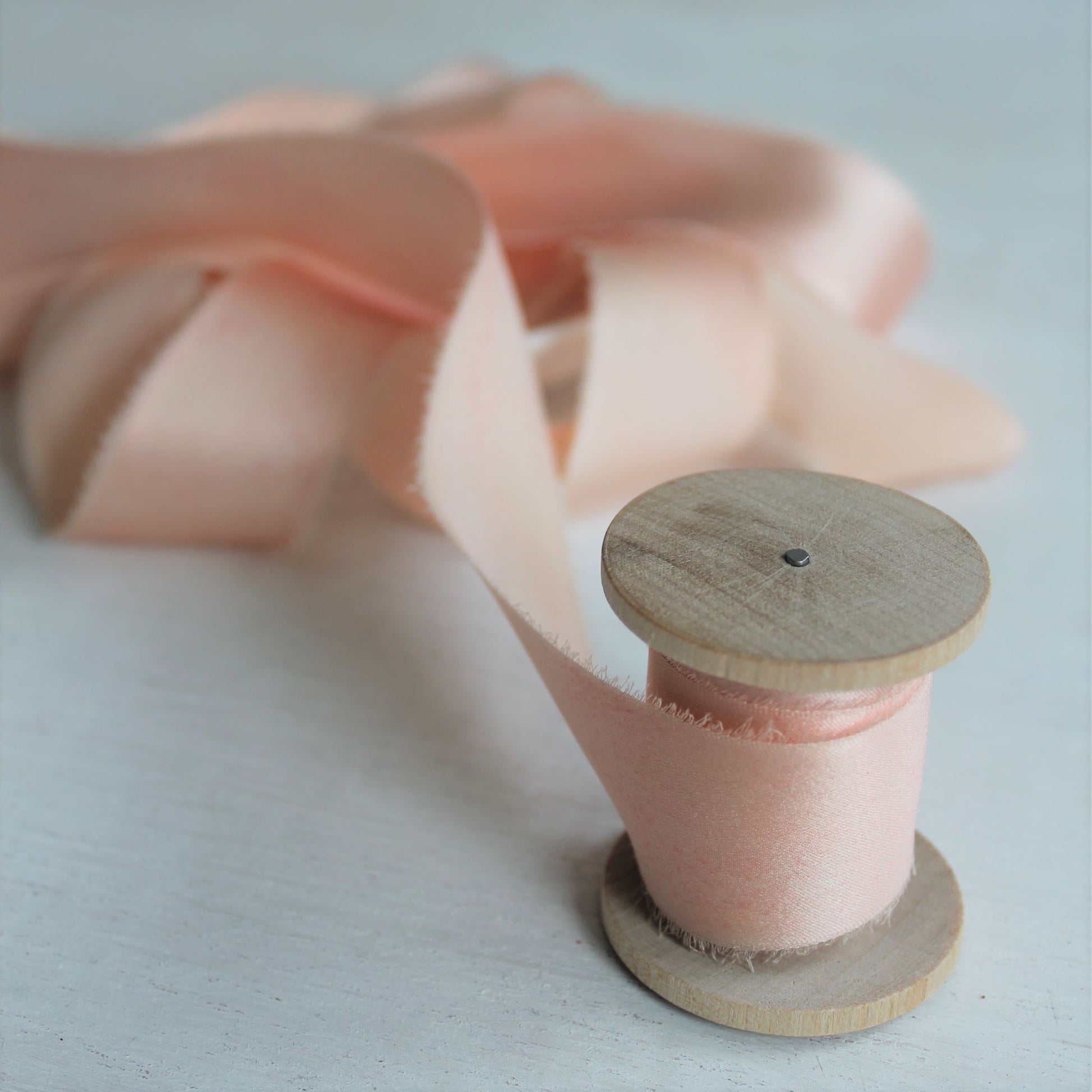 Soft, hand-torn pink blush silk charmeuse ribbon with delicate feathered edges on a wooden spool.